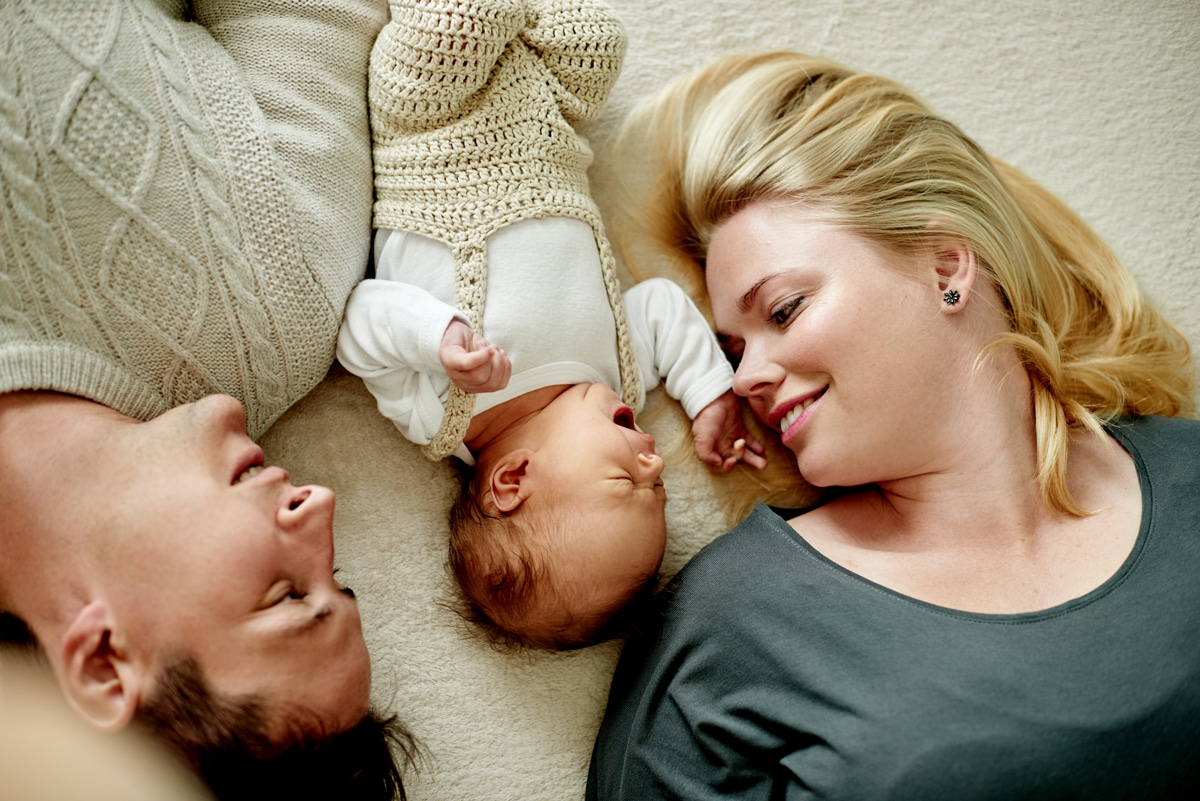 junge Mutter liegt mit jungem Vater und Baby dazwischen auf Teppichboden