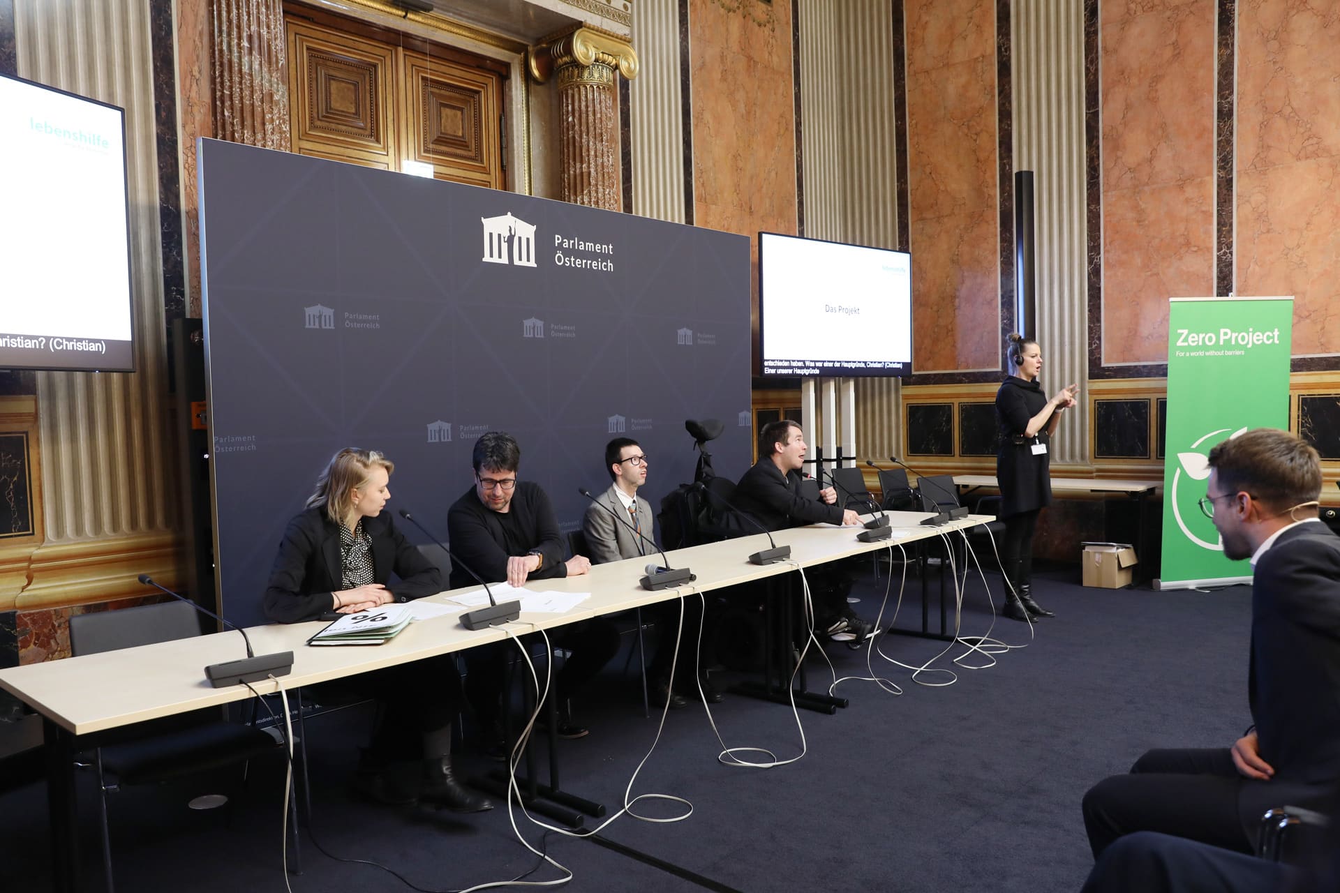 Drei Männer, zwei davon mit Behinderung und eine Frau sitzen im Anzug im österreichischen Parlament und halten Rede
