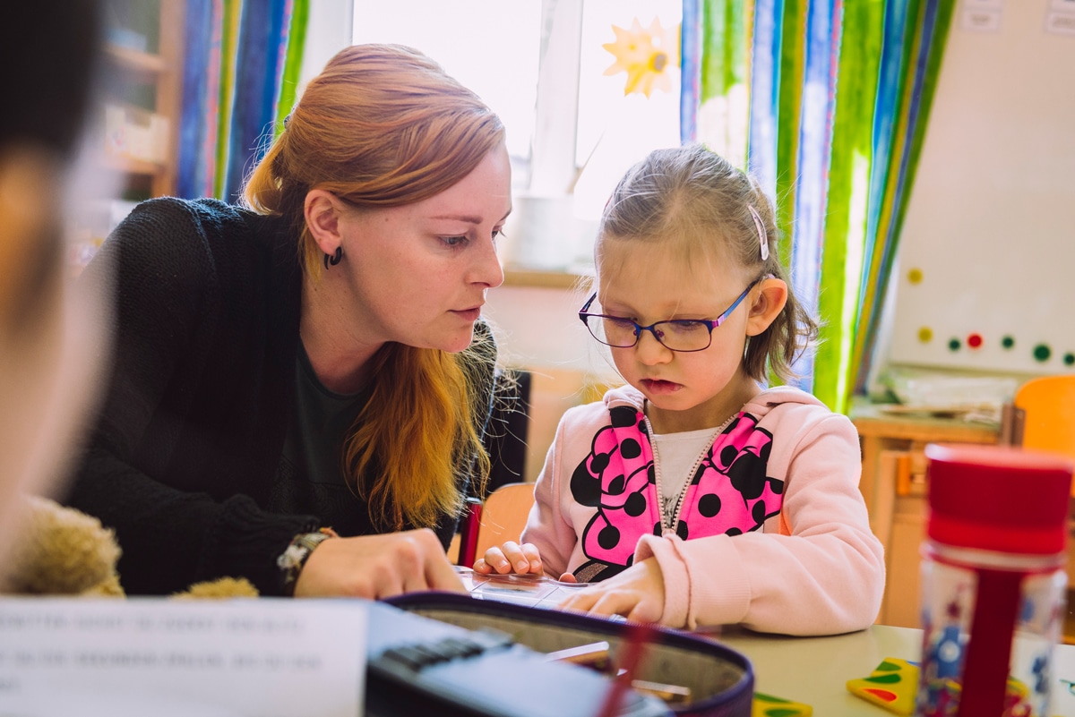 Schulassistentin unterstützt kleines Mädchen mit Brille beim Lernen