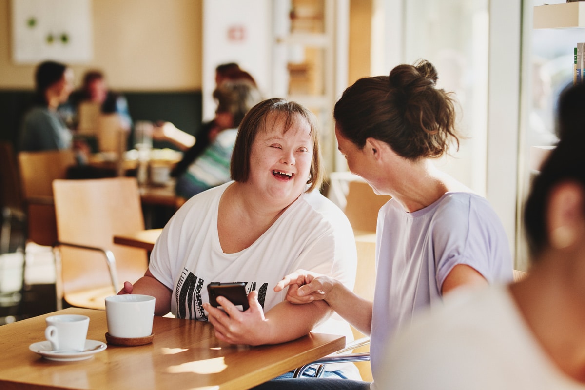 Frau mit Downsyndrom mit Freizeitassistentin sitzen lachend in einem Kaffee und reden miteinander