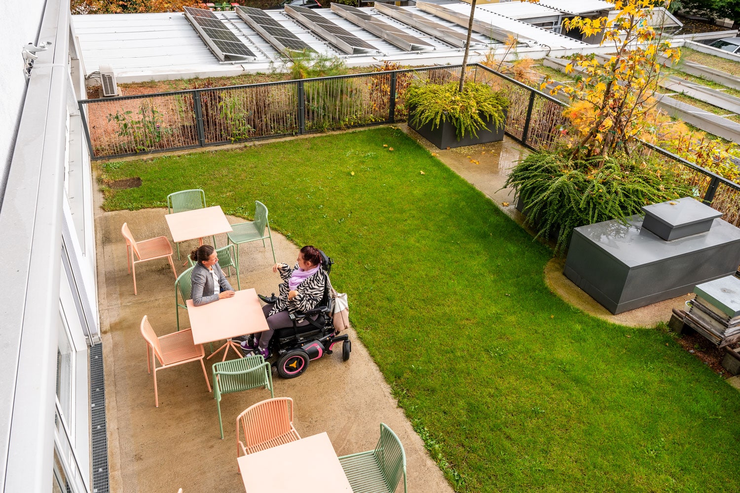 Frau im Rollstuhl und Dame an einem Tisch sitzend auf Dachterrasse mit PV Anlage und Wiese