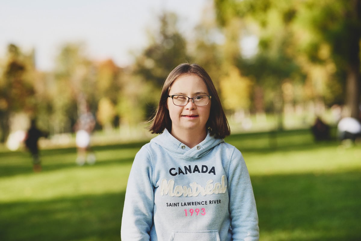 junges Mädchen mit Downsyndrom und Brille steht in Park und blickt selbstbewusst in die Kamera