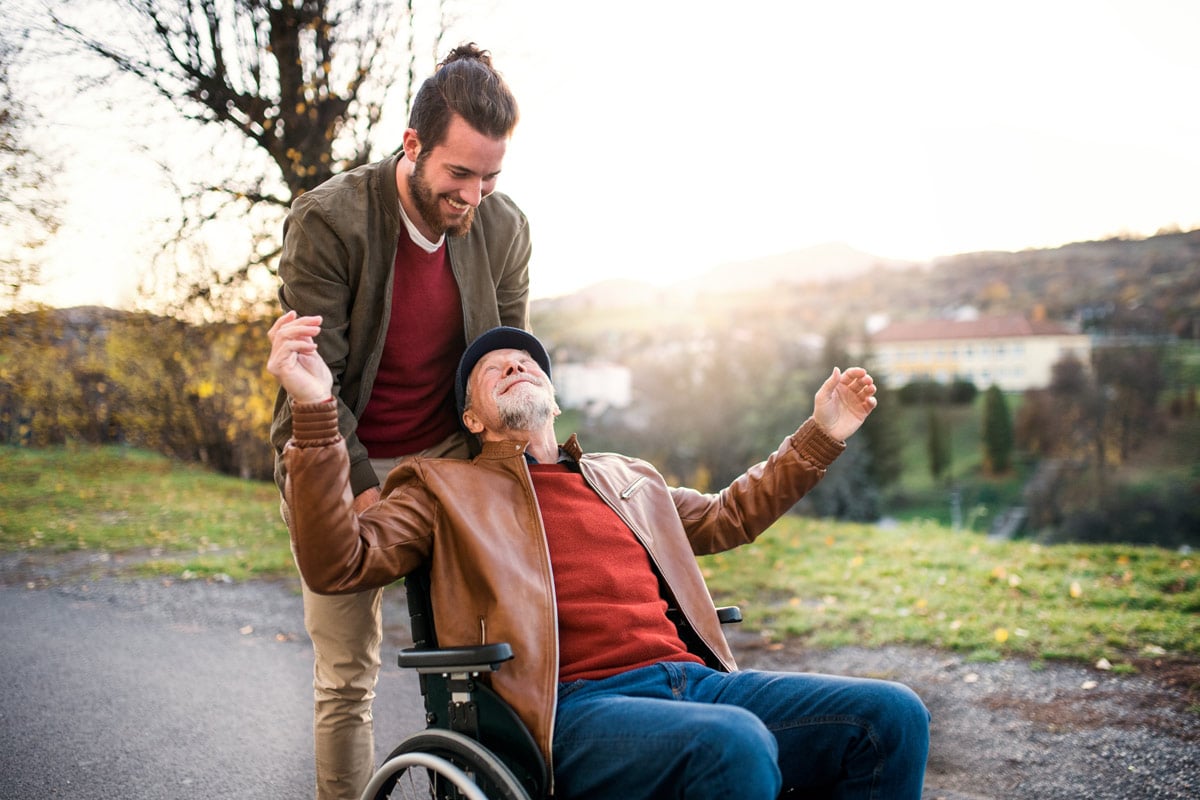 Mann schiebt älteren Herren im Rollstuhl an und sie haben viel Spaß dabei