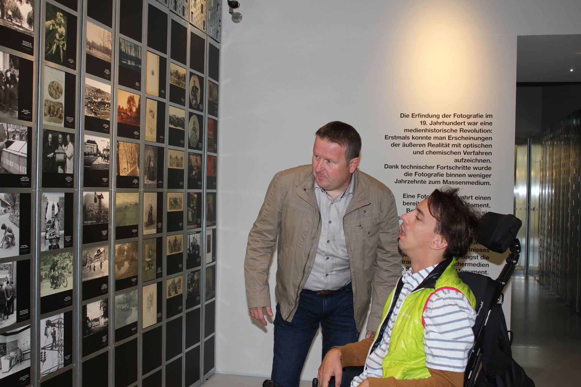 Mann im Rollstuhl und Begleiter betrachten eine Fotoausstellung der Initiative Kultur Inklusiv im Museum