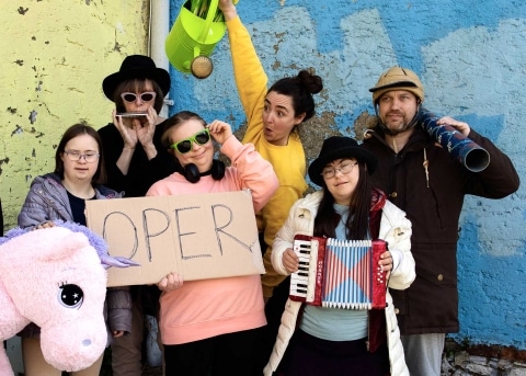 Auf dem Bild ist eine Gruppe von Menschen mit und ohne Behinderungen, die kostümiert sind, und eine Person hält ein Schild mit der Aufschrift Oper