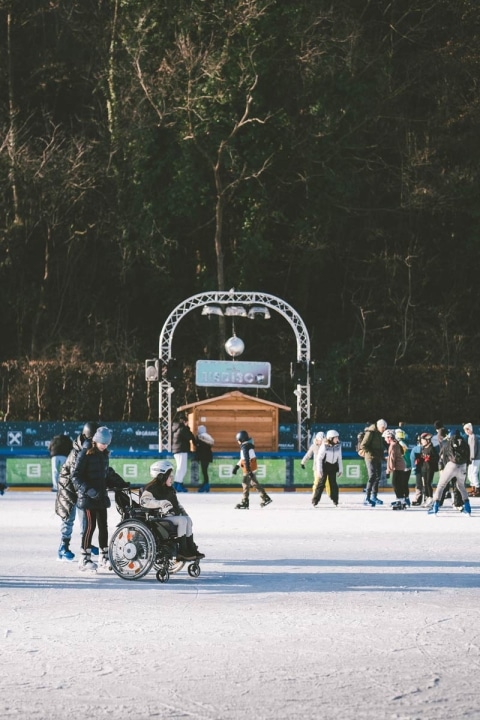 Eine Frau in Schlittschuhe läuft mit einer Rollstuhlfahrerin auf einem Eislaufplatz