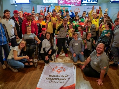 Menschegruppe von mehr als 40 Personen stehend bzw. in Hocke vor Special Olympics Fahne, die am Boden gehalten wird