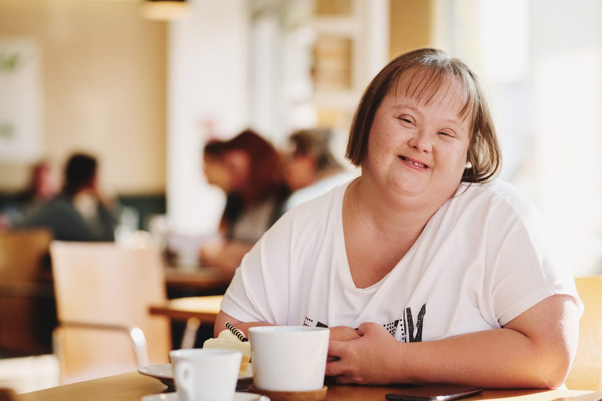 junge Frau mit Down Syndrom sitzt in einem Café und trinkt Cappuchino
